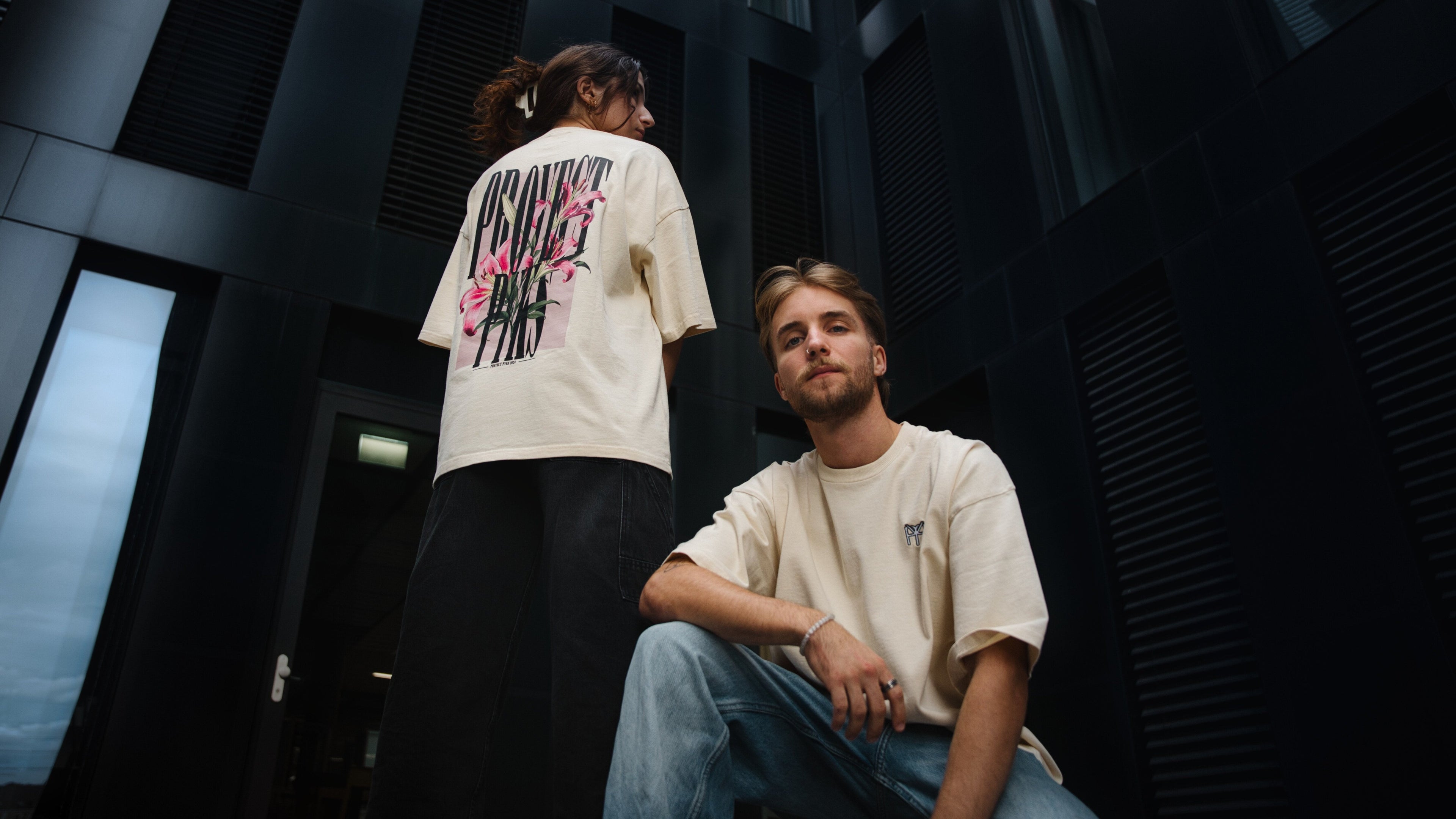 Beiges oversized T-Shirt 'Desert Flora' mit detailliertem Pflanzenmotiv auf der Hinterseite – modernes und urbanes Design für lässigen Stil, Bild von Vorne, Model männlich, Model weiblich Logo in weiss mit schwarzem Rand auf der Vorderseite, schwarzes Gebäude als Hintergrund